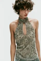 Top halter confeccionado en mezcla de poliamida, color verde militar, con diseño cut-out en el escote, espalda descubierta y aplicaciones de abalorios y lentejuelas brillantes. Presenta bajo recto con flecos colgantes de cuentas y cierre posterior con botones en el cuello.