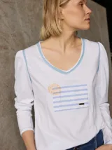 Remera de manga larga blanca con escote en V y vivos en color celeste. Presenta un bolsillo frontal con estampado de rayas celestes y un diseño circular bordado en color amarillo. Las mangas tienen un ligero fruncido en los hombros.
