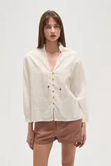 Camisa blanca de manga larga, con cuello mao y escote en V ajustable mediante cordones terminados en mostacillas decorativas.