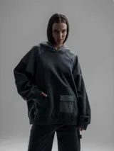 Hoodie oversize de color gris oscuro, confeccionado en frisa de algodón con recortes en textiles técnicos. Presenta capucha forrada, bolsillo frontal tipo canguro y un bolsillo adicional con solapa en la parte inferior.