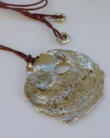 Collar artesanal con dije central de metal con textura orgánica irregular que evoca una ostra. El dije está montado sobre un cordón de algodón natural, permitiendo un uso versátil y regulable.