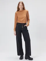 Jeans de corte ancho (wide leg) y tiro medio, confeccionados en denim de algodón negro con costuras contrastantes en color ocre. Presenta múltiples botones metálicos frontales y tachas metálicas aplicadas a lo largo de los laterales y la pretina.