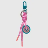 Llavero decorativo con mosquetón metálico color petróleo, argolla plateada y cordón trenzado bicolor en rosa y blanco. Incluye un dije circular de goma con el logo de la marca grabado.
