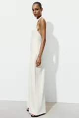 Mono largo de color blanco con cuello halter y espalda descubierta. Presenta un diseño de pierna ancha y cierre posterior con cremallera oculta y botones.