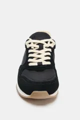 Zapatilla deportiva tipo running con diseño de paneles en contraste. Presenta cuerpo en color negro con refuerzos de gamuza sintética, detalle de pespuntes, talón en color crema y suela dentada bicolor.