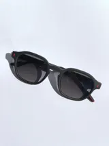 Lentes de sol con armazón de marco completo en color gris mate, con detalles decorativos en color rojo en las esquinas y patillas. Cuentan con cristales oscuros polarizados con protección UV 400.