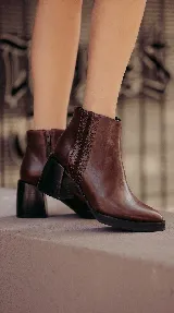 Bota corta de cuero color marrón con acabado liso, que presenta un detalle de textura acanalada en el lateral y cierre interno. Cuenta con un taco ancho y estable de color negro.