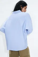 Camisa de manga larga color celeste claro, confeccionada en popelín de algodón. Presenta cuello solapa con botones y un bolsillo de parche en el pecho izquierdo.