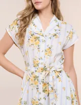 Vestido camisero midi de silueta evasé, confeccionado en tejido liviano blanco con estampado floral amarillo y rayas celestes. Cuenta con cuello solapa en V, cartera con botones frontales, mangas cortas japonesas y cintura definida con lazo ajustable a juego.