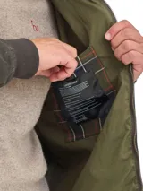 Campera de algodón encerado en color verde oliva, con cierre frontal, puños y ruedo elásticos, y cuello de pana en color marrón con detalle de broches metálicos.