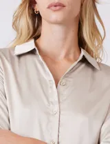 Camisa de satén color verde oliva, con cuello clásico, cierre frontal con botones metálicos y mangas largas con puños abotonados.