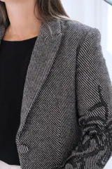 Blazer de corte sastrero en color gris con patrón de rayas diagonales y bordados negros en las mangas y solapas. Presenta cierre frontal con botón, bolsillos con solapa y estructura clásica.