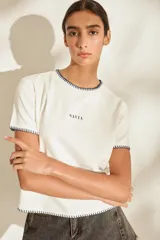 Remera de manga corta color crudo, con detalles de costuras decorativas en contraste en cuello, puños y dobladillo. Presenta el nombre de la marca impreso en el centro del pecho.