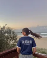 Camiseta de fútbol de Escocia de manga corta, color azul marino con diseño a cuadros sutiles, cuello en V y cuello camisero blanco con líneas azules. Presenta detalles estampados en el frente, incluyendo el escudo de la Asociación de Fútbol de Escocia, un león rampante y el texto 'Scottish Football Association'.