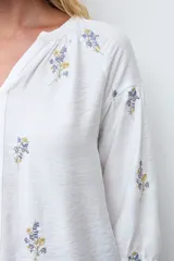 Blusa blanca de corte holgado con escote en V y mangas tres cuartos abullonadas. Presenta un delicado bordado de pequeñas flores en tonos azules y amarillos dispersas por toda la prenda.