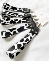 Llavero de muñeca con cinta de tela estampada con diseño de vaca en blanco y negro. Tiene un conector de cuero sintético negro con remaches plateados y un gancho metálico plateado.
