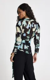 Blusa de tejido fluido con estampado abstracto en tonos azules, negros y beige. Presenta un diseño cruzado con escote en V, mangas 3/4 anchas y un lazo lateral ajustable que define la silueta.