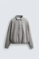 Sudadera de corte boxy fit en color gris, confeccionada en tejido de algodón con interior afelpado. Presenta cuello solapa, cierre frontal con botones a presión, manga larga con puños acanalados y bolsillo frontal tipo canguro. El bajo cuenta con un acabado elástico.