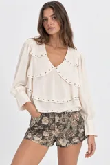 Blusa de seda color marfil con escote en V, mangas largas con puños fruncidos y múltiples volados en capas adornados con tachas metálicas.