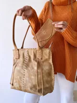Cartera tipo tote de cuero color camel, con diseño artesanal que presenta costuras decorativas visibles en los laterales y el frente. Cuenta con doble asa de hombro y cierre superior.
