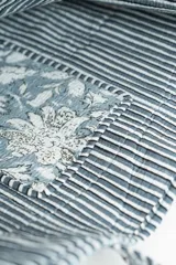 Bolso tipo tote bag de tela acolchada con estampado floral en tonos gris y blanco. Presenta asas largas reforzadas con detalle de rayas en los bordes.