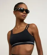 Top deportivo negro, corte tipo bralette, con escote asimétrico y doble tirante en los hombros. Presenta una banda elástica inferior con una delgada línea blanca horizontal.