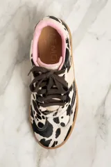 Zapatillas bajas de cuero con estampado de vaca en blanco y negro, cordones marrones y suela de goma color caramelo.