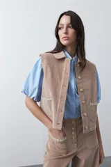 Chaleco de pana color marrón oscuro, con cuello camisero, cierre frontal de botones metálicos dorados y dos bolsillos frontales con detalle de bordado geométrico.