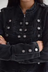 Chaqueta de estilo militar confeccionada en denim negro con efecto lavado. Presenta un diseño estructurado con cierre frontal de cremallera, cuello mao y múltiples botones metálicos decorativos dispuestos en filas horizontales. Incluye detalles de charreteras en los hombros y puños con botones.