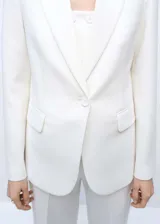Chaqueta tipo americana de diseño estructurado y corte recto, color blanco. Presenta cuello con solapas de muesca, cierre frontal con un botón, mangas largas con puños abotonados y dos bolsillos de parche frontales. Incluye forro interior.