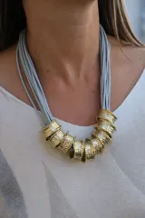 Collar de diseño artesanal compuesto por múltiples tiras de cuero gamuzado en color gris claro, adornado con una serie de piezas cilíndricas de metal dorado con textura martillada.