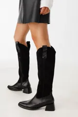 Botas bucaneras negras de cuero y gamuza, con taco de 5cm, punta cuadrada y caña alta. Cuentan con un tirador en la parte superior de la caña.