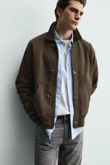 Chaqueta de algodón con interior de rizo, color marrón con efecto lavado. Presenta cuello de pana, bolsillos frontales tipo canguro, puños y ruedo en rib, y cierre frontal con alamares metálicos.