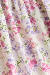 Vestido corto de algodón con estampado floreado en tonos rosados y lilas sobre base blanca. Presenta escote en V fruncido, mangas globo a los codos con puños elásticos y sección fruncida en la cintura.