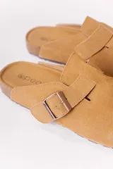 Zueco de gamuza color tostado, con diseño cerrado en la parte delantera, tira ajustable con hebilla metálica en el empeine, plantilla anatómica y suela tipo corcho.