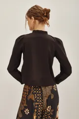 Camisa blanca de algodón con cuello alto, cierre frontal de botones ocultos y detalles de bordado floral en el pecho. Presenta mangas largas con puños elásticos y volados decorativos en la parte frontal.