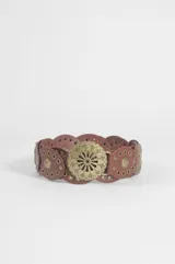 Cinturón de cuero vegano color marrón, compuesto por una serie de piezas circulares unidas entre sí, cada una con un aplique metálico central en forma de sol y detalles de tachas circulares. Incluye una hebilla circular grande con diseño grabado.