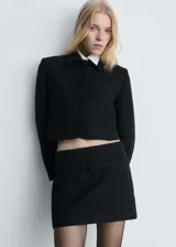 Chaqueta corta de tejido tweed en color negro, con cuello camisero, manga larga y cierre frontal con botones forrados. Presenta un diseño recto con dos bolsillos laterales.