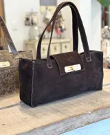 Cartera tipo tote bag de formato rectangular y alargado, confeccionada en gamuza color marrón oscuro. Presenta doble asa de hombro, solapa frontal con cierre metálico giratorio en tono dorado y costuras visibles en los bordes.