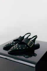 Chatas de cuero negro con punta fina, decoradas con tachas metálicas plateadas. Presentan una tira con hebilla ajustable sobre el empeine y diseño destalonado con tira trasera.