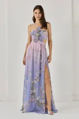 Vestido largo de gasa color lila con estampado floral, escote halter y detalle de gran flor en 3D en el pecho. Cuenta con aberturas laterales en la cintura y falda amplia con tajo pronunciado en la pierna.