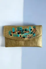 Sobre de mano y monedero de cuero dorado con solapa y arte pintado a mano en tonos turquesa y negro. Cuenta con dos compartimentos internos y cierre con imán.