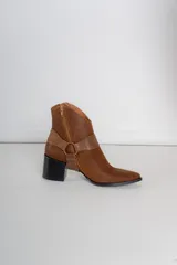 Bota corta de cuero color bordó, con taco cuadrado y punta fina. Presenta una correa con tachas pequeñas y una argolla metálica decorativa alrededor del tobillo. Cuenta con cierre lateral.