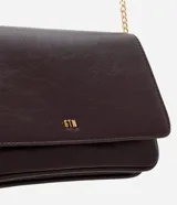 Cartera bandolera pequeña de poliuretano color marrón oscuro con acabado texturizado. Cuenta con solapa frontal, logo metálico dorado en la parte inferior y correa larga de cadena metálica dorada.