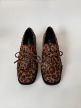 Mocasín de cuero marrón con textura granulada, diseño de horma cuadrada, detalle de flecos y lazo decorativo en el empeine, montado sobre una suela y taco bajo de color negro.