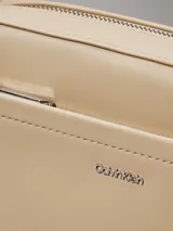 Cartera rectangular tipo bandolera de piel sintética color beige claro, con bolsillo exterior frontal y cierre superior de cremallera. Incluye dos correas desmontables: una ancha y una fina ajustable. Presenta el logo metálico de Calvin Klein en la parte frontal.
