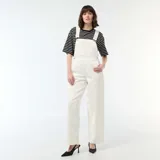 Enterito de jean color blanco con diseño de pierna ancha, bolsillo frontal en el pecho y tirantes ajustables con hebillas metálicas.