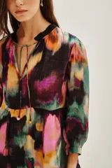 Vestido corto con estampado abstracto en tonos vibrantes como fucsia, verde, amarillo y negro. Presenta cuello con lazo ajustable y mangas largas abullonadas con puños elásticos. El cuerpo tiene un corte imperio con un volante fruncido en el ruedo.
