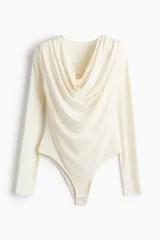 Body de manga larga en punto elástico color crema, con escote drapeado y hombreras. Presenta costuras fruncidas para realzar el efecto drapeado, refuerzo interior con botones a presión y cobertura media en la parte posterior.