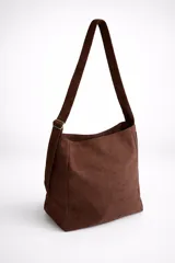 Bolso tipo bandolera de tamaño mediano, confeccionado en material con acabado efecto gamuza color chocolate. Presenta un diseño minimalista con cierre superior de cremallera y correa larga regulable con herrajes metálicos.